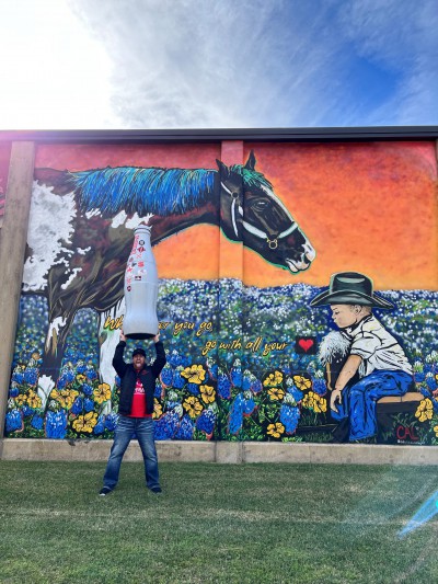 Blake Smith, Distribution Supervisor, hoists the Recognition Roadtrip Bottle in front of a cowboy themed mural in downtown Bryan that offers the advice, “Wherever you go, go with all your heart.”